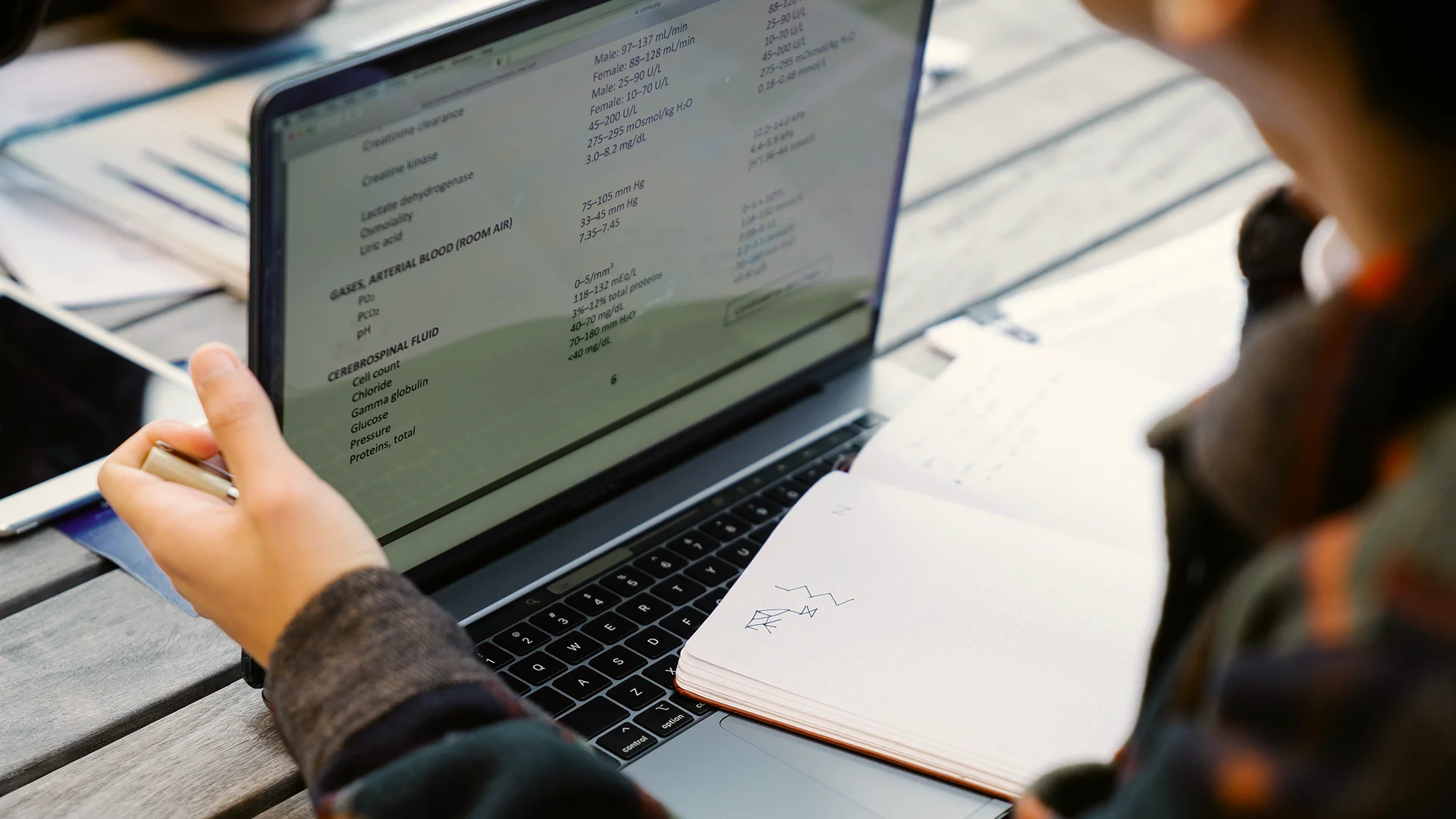 Student working on a laptop with an open document while taking notes in a notebook