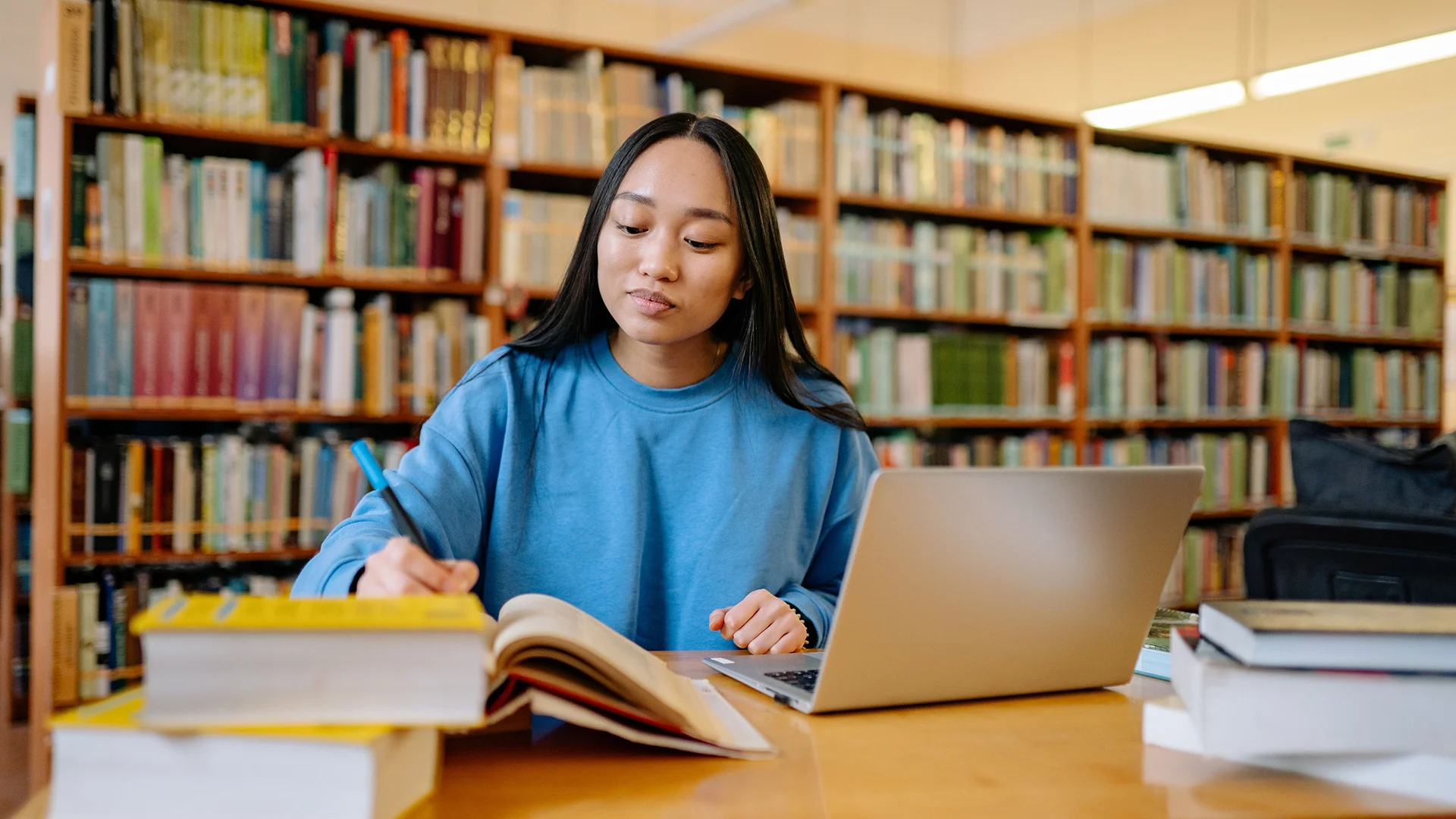 Student studying in a library with a laptop, open book and handwritten notes