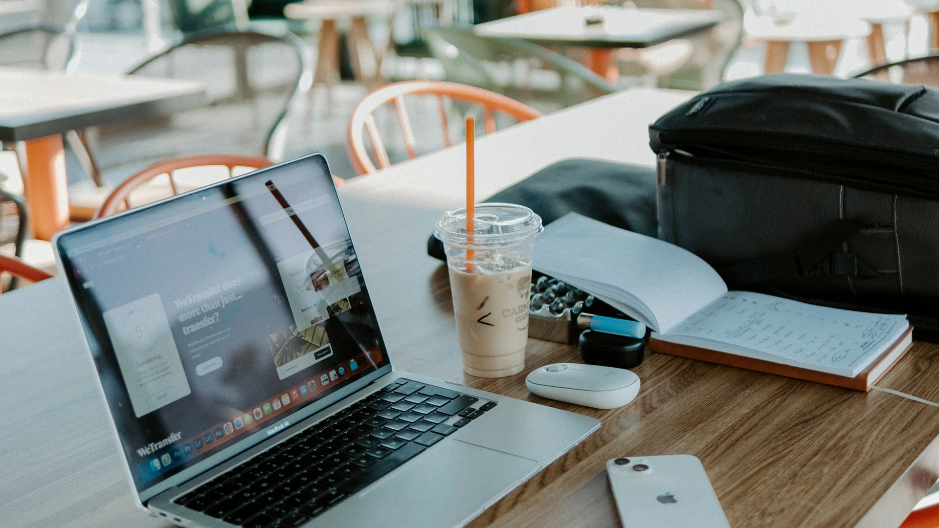 Laptop on a table with a mouse, phone, notebook, pen, earphones and a drink