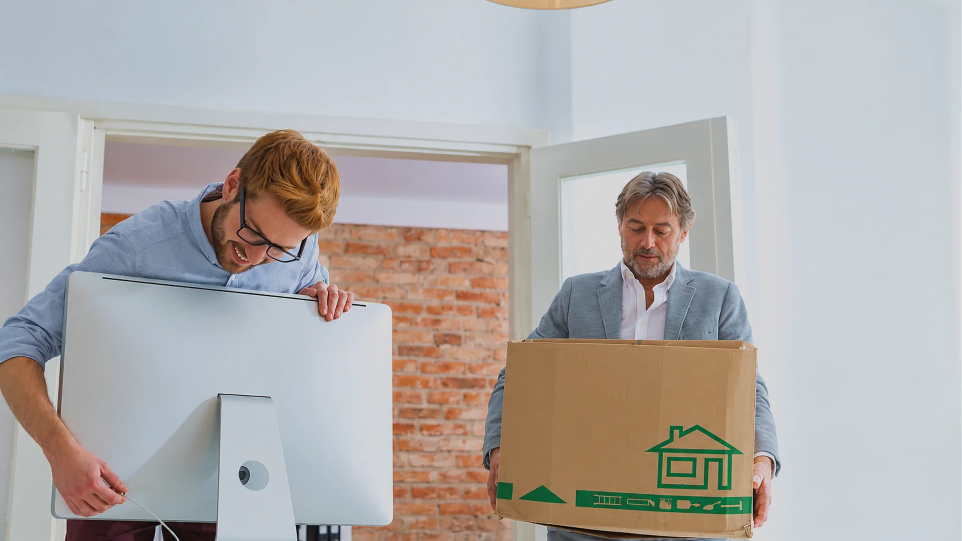 Two men move computer equipment: one holds a monitor and disconnects a cable, while the other carries a cardboard box